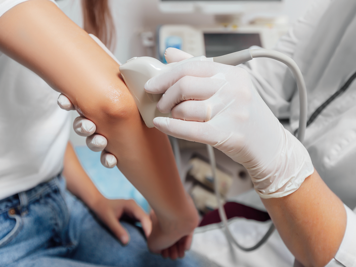 Arzt führt Ultraschalluntersuchung durch. Er führt den Schallkopf des Ultraschallgerätes am Ellenbogen der Patientin entlang.