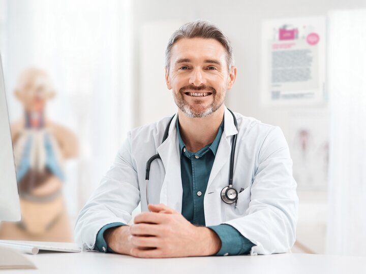 Arzt mit Dreitagebart, Arztkittel und Stethoskop sitzt lächelnd am Schreibtisch. Dahinter verschwommen ein Plakat sowie eine Anatomiefigur.