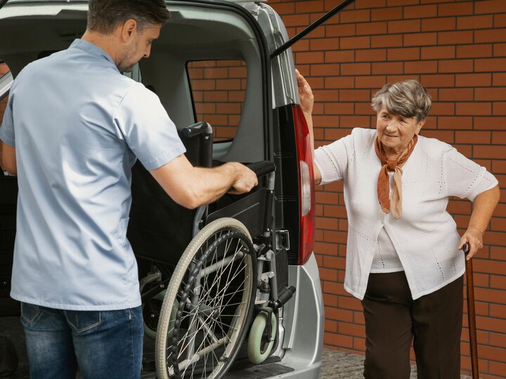 Betreuer holt Rollstuhl aus dem Auto für gehbehinderte ältere Frau mit Gehstock